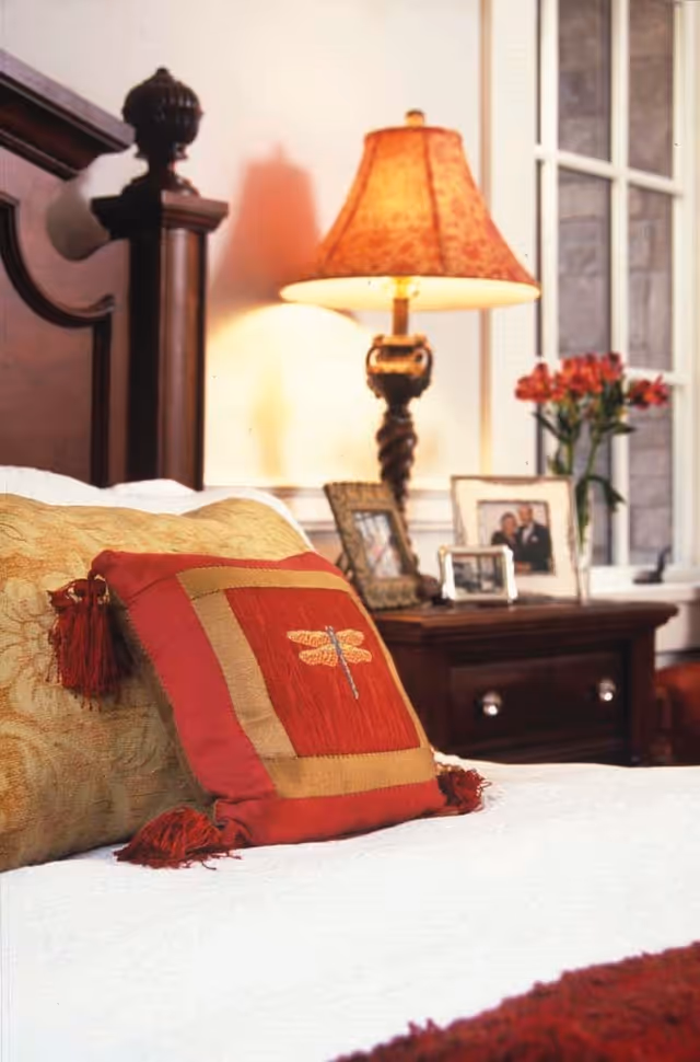 Close-up view of a bed with decorative pillows, including a red pillow with a dragonfly design, in a warmly lit bedroom. In the background, there is a wooden nightstand with framed photos, a table lamp with a patterned shade, and a vase with red flowers near a window.
