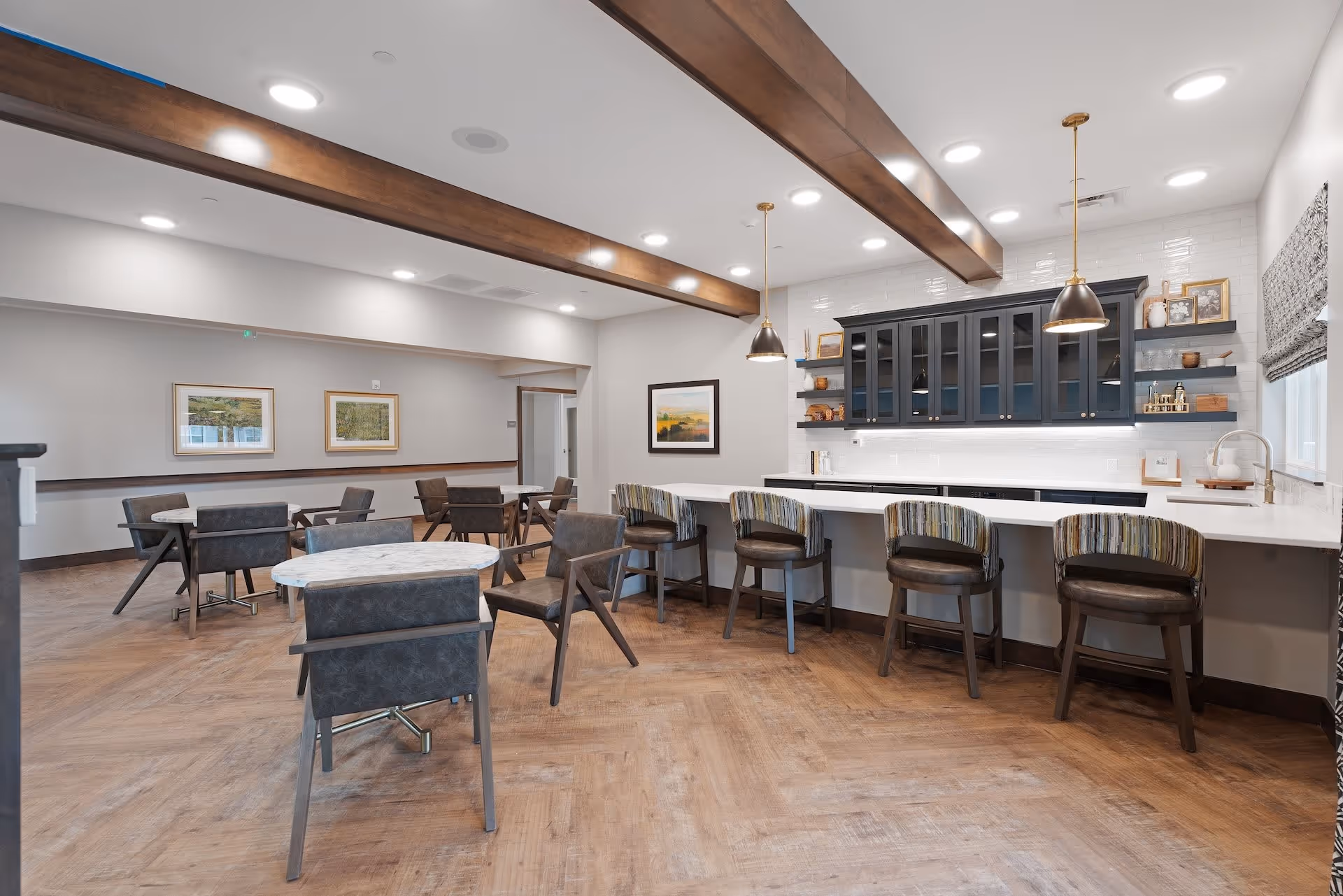Bright communal dining area with a long marble-topped bar and striped barstools, round tables and chairs, and exposed wooden ceiling beams.