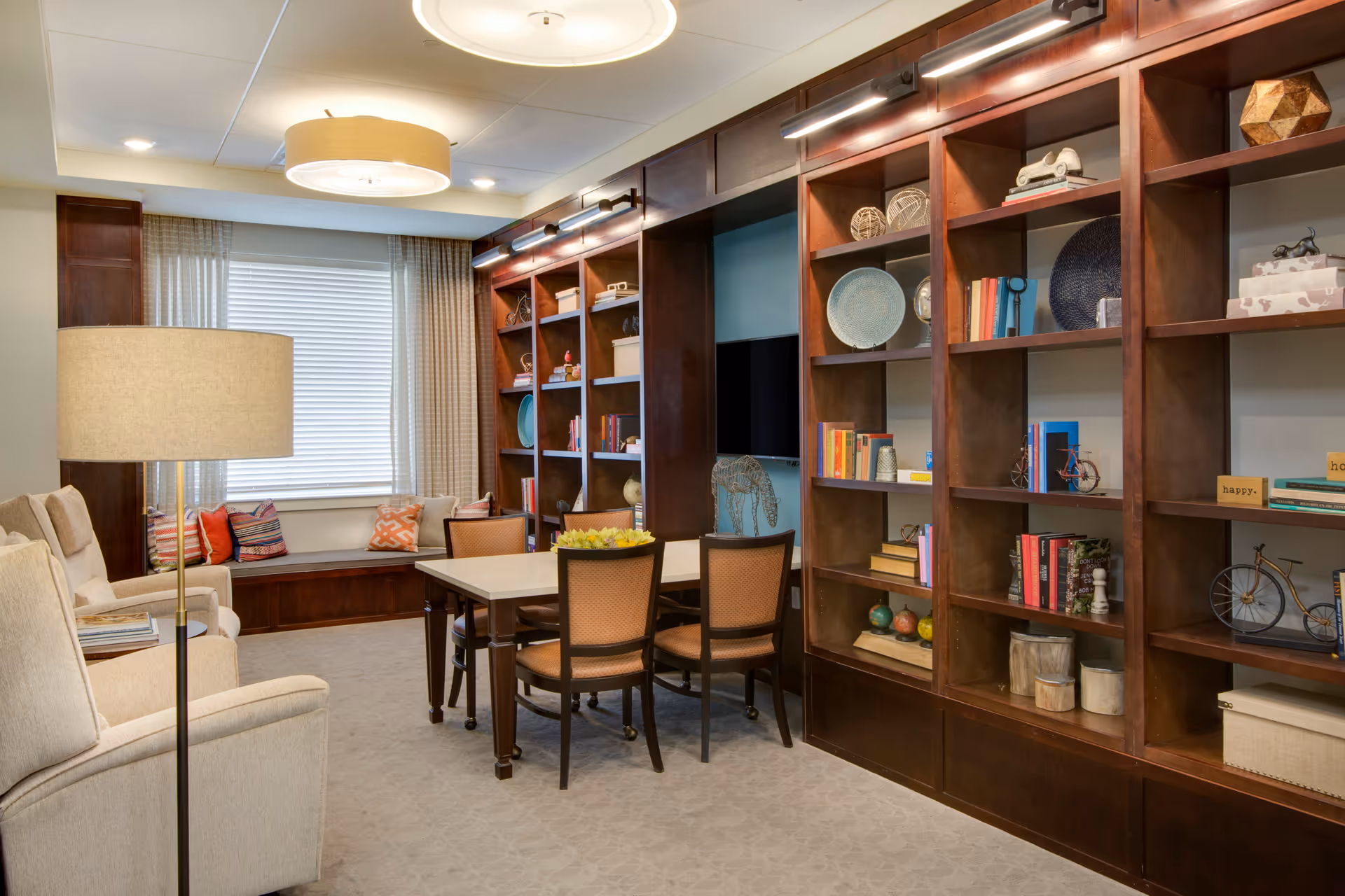 A cozy living room area with beige armchairs, a floor lamp, and a wooden table surrounded by four chairs. The room features large wooden bookshelves filled with decorative items, books, and art pieces. A window with beige curtains and a cushioned window seat with colorful pillows is visible in the background.