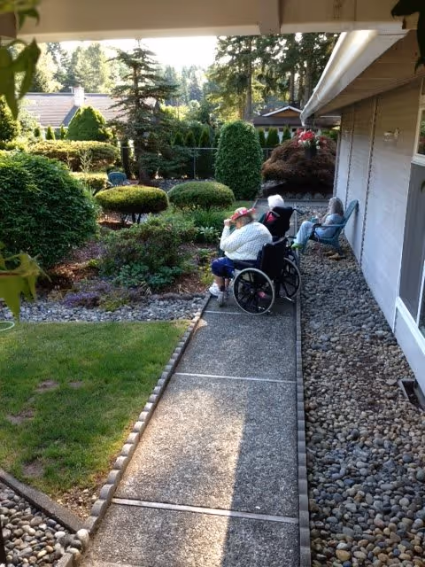 Two elderly individuals sitting outside along a paved pathway next to a building. One person is in a wheelchair wearing a hat, and the other is seated in a chair. The area is landscaped with bushes, trees, and a lawn under a sunny sky.