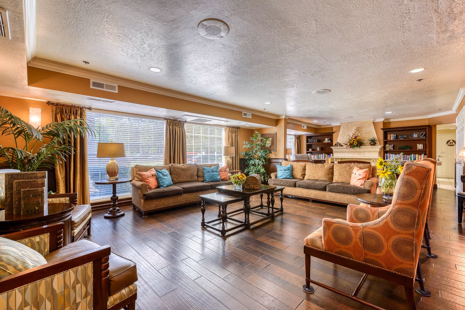 A spacious and well-lit living room in a senior living facility featuring two large sofas with colorful cushions, two patterned armchairs, a wooden coffee table, and a fireplace with decorative items. Large windows with curtains allow natural light to fill the room, and there are plants and bookshelves adding to the cozy atmosphere.