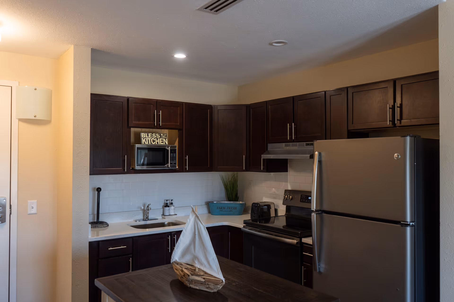 A modern kitchen with dark wood cabinets, a stainless steel refrigerator, stove, and microwave. The countertop has a small sink, soap dispensers, a toaster, and a decorative plant. A small wooden table with a decorative sailboat is in the foreground.