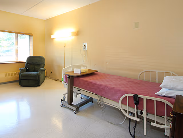 A simple, clean hospital-style bedroom with a single bed covered in a pink blanket, a small overbed table, a green recliner chair near a window with blinds, and a wall-mounted light fixture illuminating the room.