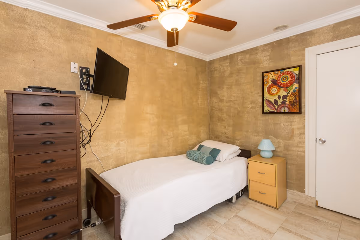 Small single-occupancy bedroom with a bed, tall dresser, wall-mounted TV, nightstand and ceiling fan.