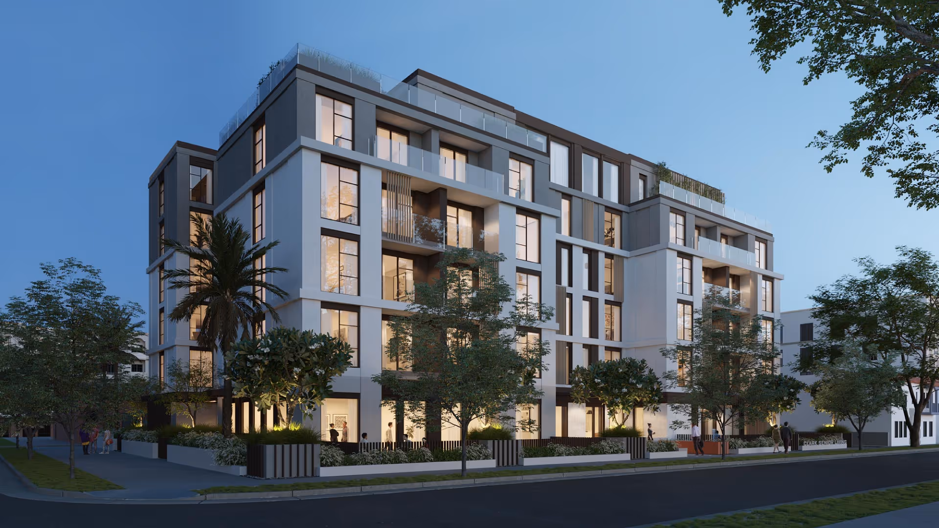 Modern multi-story senior living facility building with large windows and balconies, surrounded by trees and landscaped greenery, viewed at dusk with interior lights glowing.