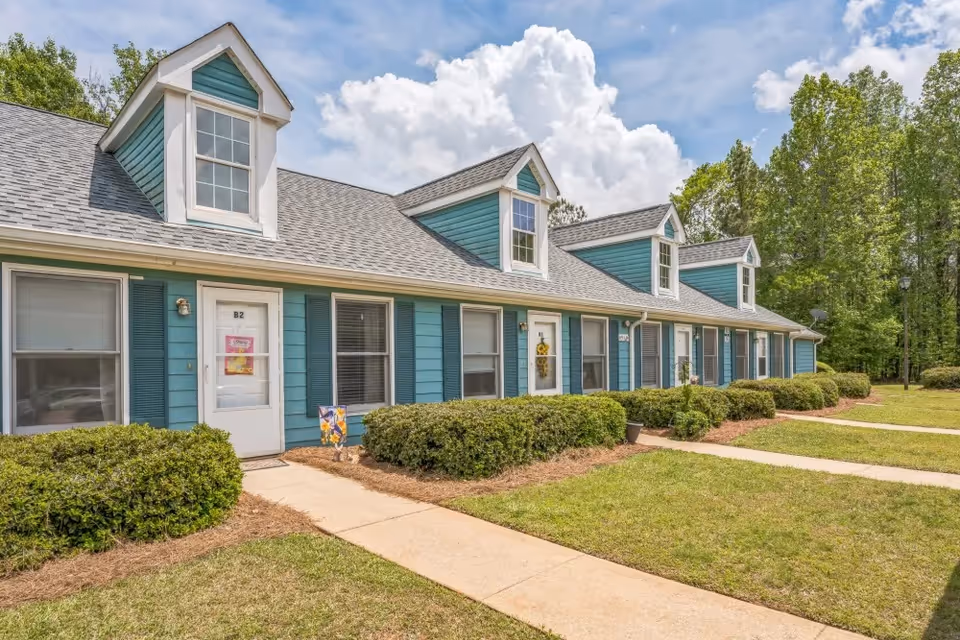 Exterior view of a single-story residential building with teal siding and white trim. The building has multiple dormer windows on the roof and several front doors, each with small bushes and a walkway leading up to them. The sky is partly cloudy and there are trees in the background.