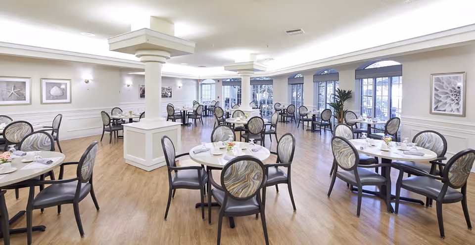 A spacious and well-lit dining room with multiple round tables, each surrounded by four chairs with patterned backs. The room features light wood flooring, white walls with wainscoting, decorative framed artwork, and large windows allowing natural light to enter. There are two large white columns in the center of the room.