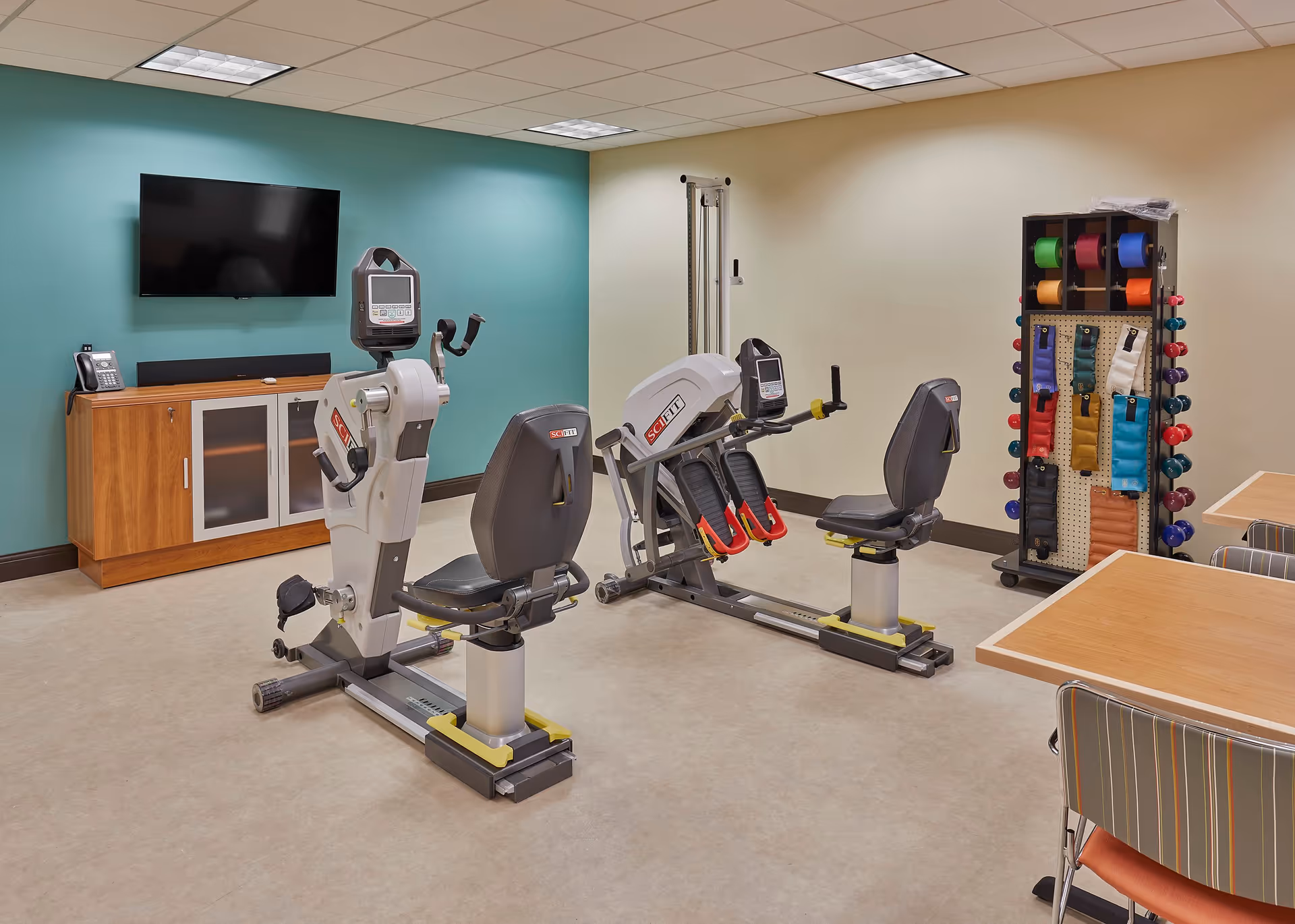 A small exercise room with two SciFit exercise machines, a wooden cabinet with a telephone and a wall-mounted TV above it, and a rack holding colorful exercise bands and small dumbbells. There are also tables and chairs with striped upholstery visible on the right side.