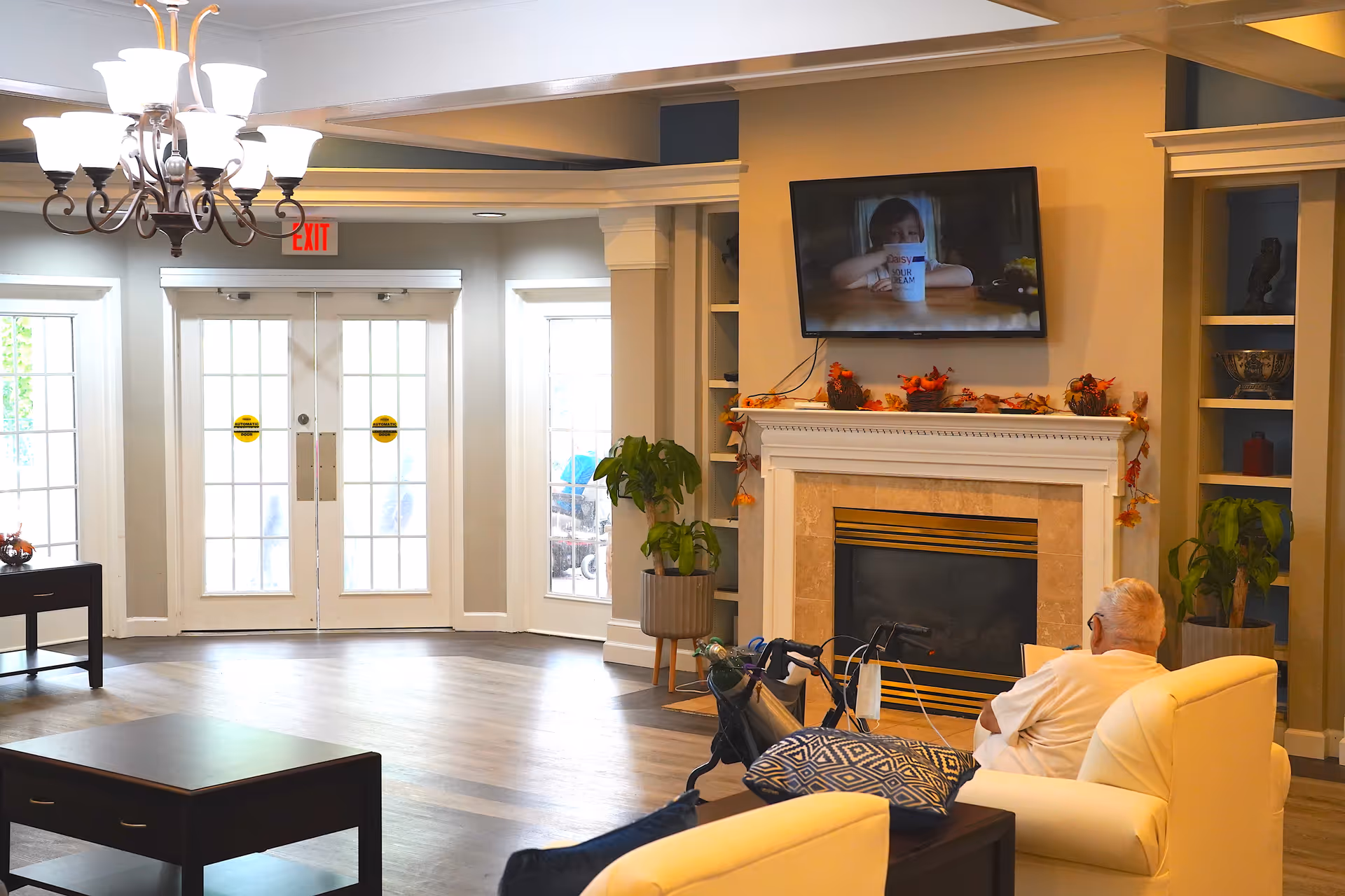 A senior man sitting in a white armchair in a cozy living room area of a senior living facility. The room features a fireplace decorated with autumn leaves and small bird figurines, a flat-screen TV mounted above the fireplace showing a child holding a cup, large windows and glass doors letting in natural light, a chandelier, and several potted plants. There is a walker with an oxygen tank next to the man.