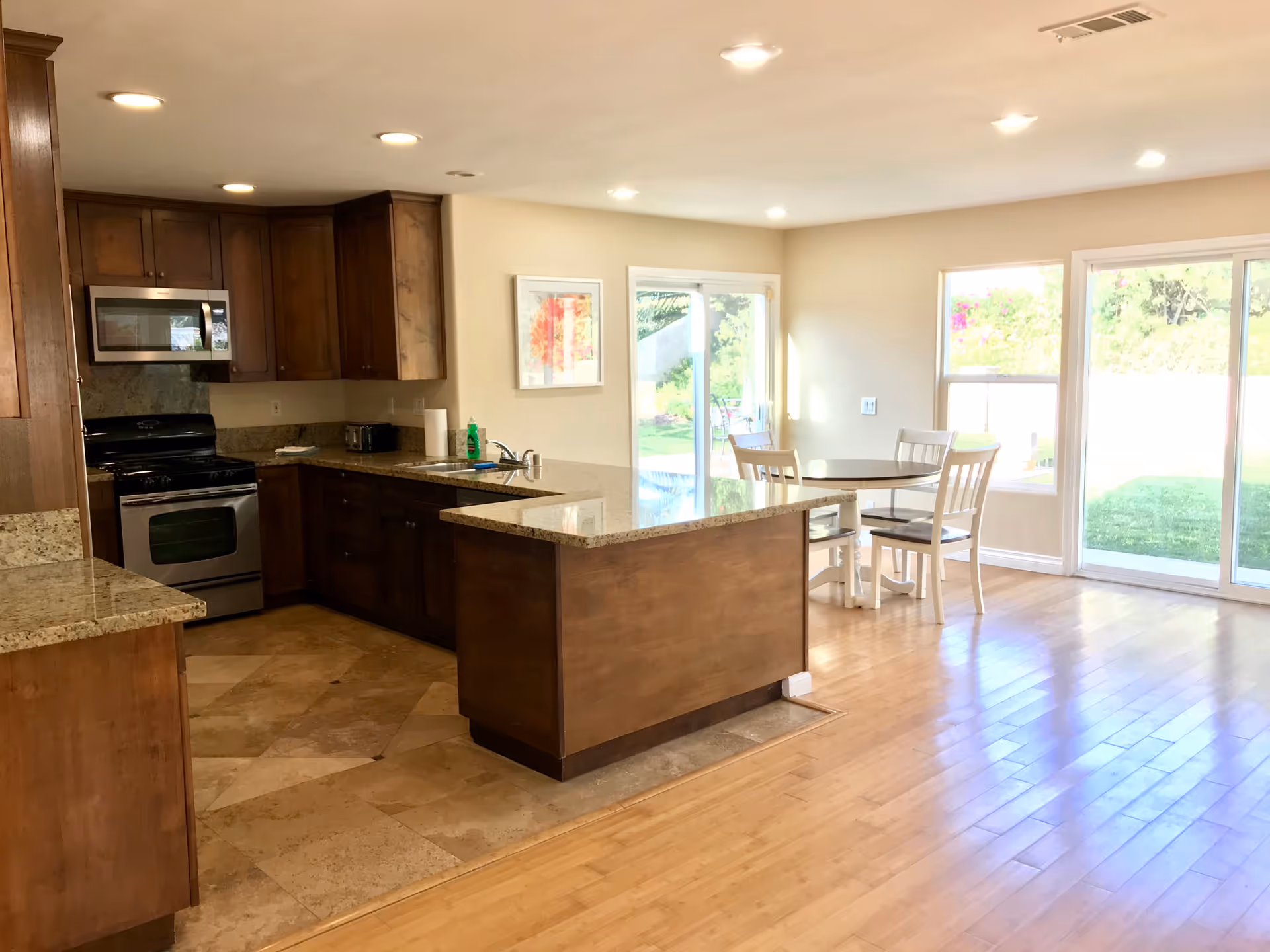 Bright kitchen and dining area with wooden cabinets, granite countertops, stainless steel microwave and oven, a small round dining table with four chairs, and large sliding glass doors leading to a green outdoor space.