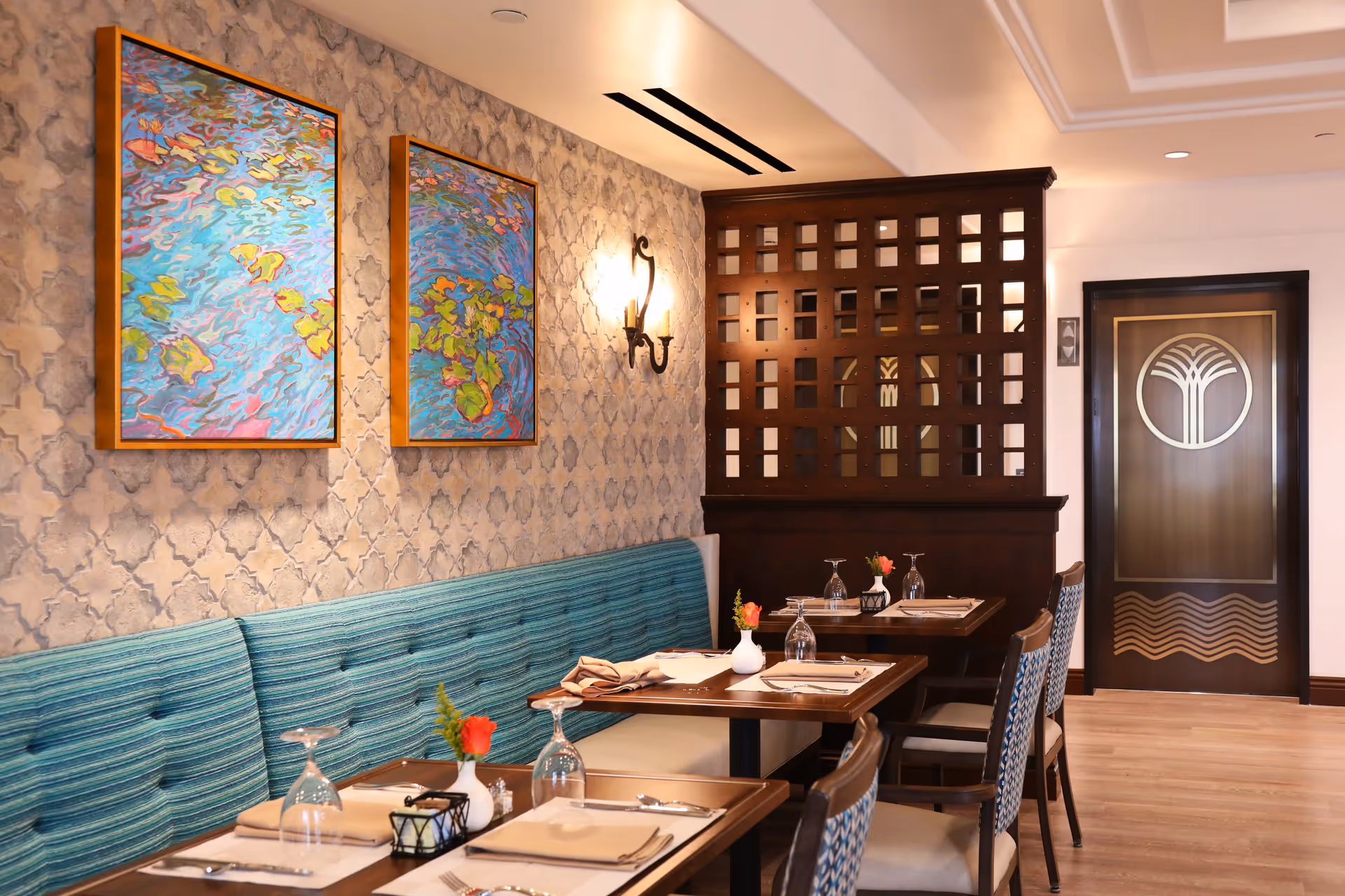 A cozy dining room with teal banquette seating, set tables with place settings and artwork on patterned wallpaper, and a wooden partition beside a decorative door.