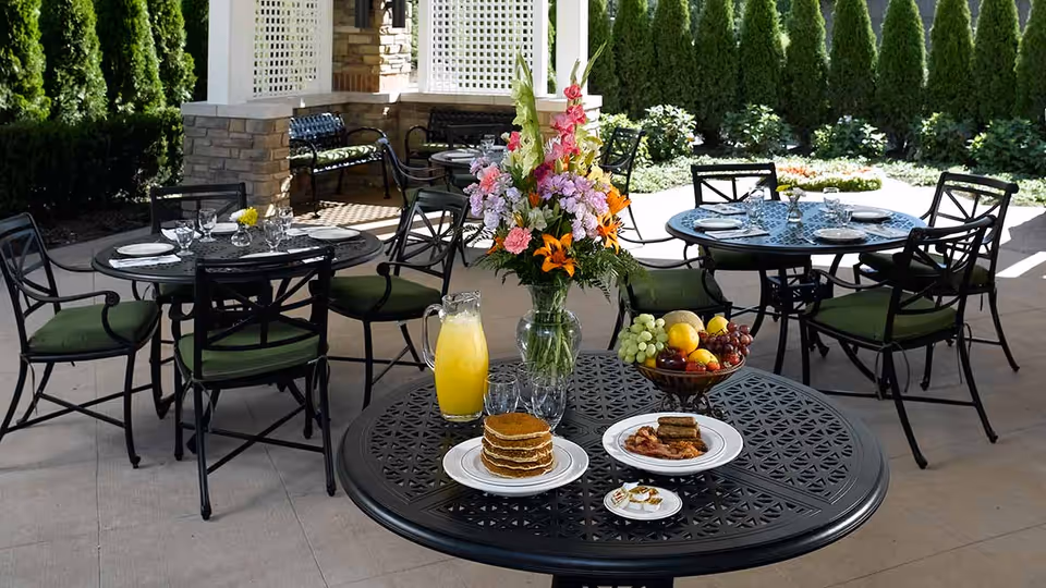 Outdoor patio area with black metal tables and chairs with green cushions. One table in the foreground has a vase with colorful flowers, a pitcher of orange juice, a plate of pancakes, a plate with bacon and sausage, a small plate with butter packets, and a bowl of assorted fruit. Other tables in the background are set with plates, glasses, and silverware. The area is surrounded by greenery and tall shrubs.