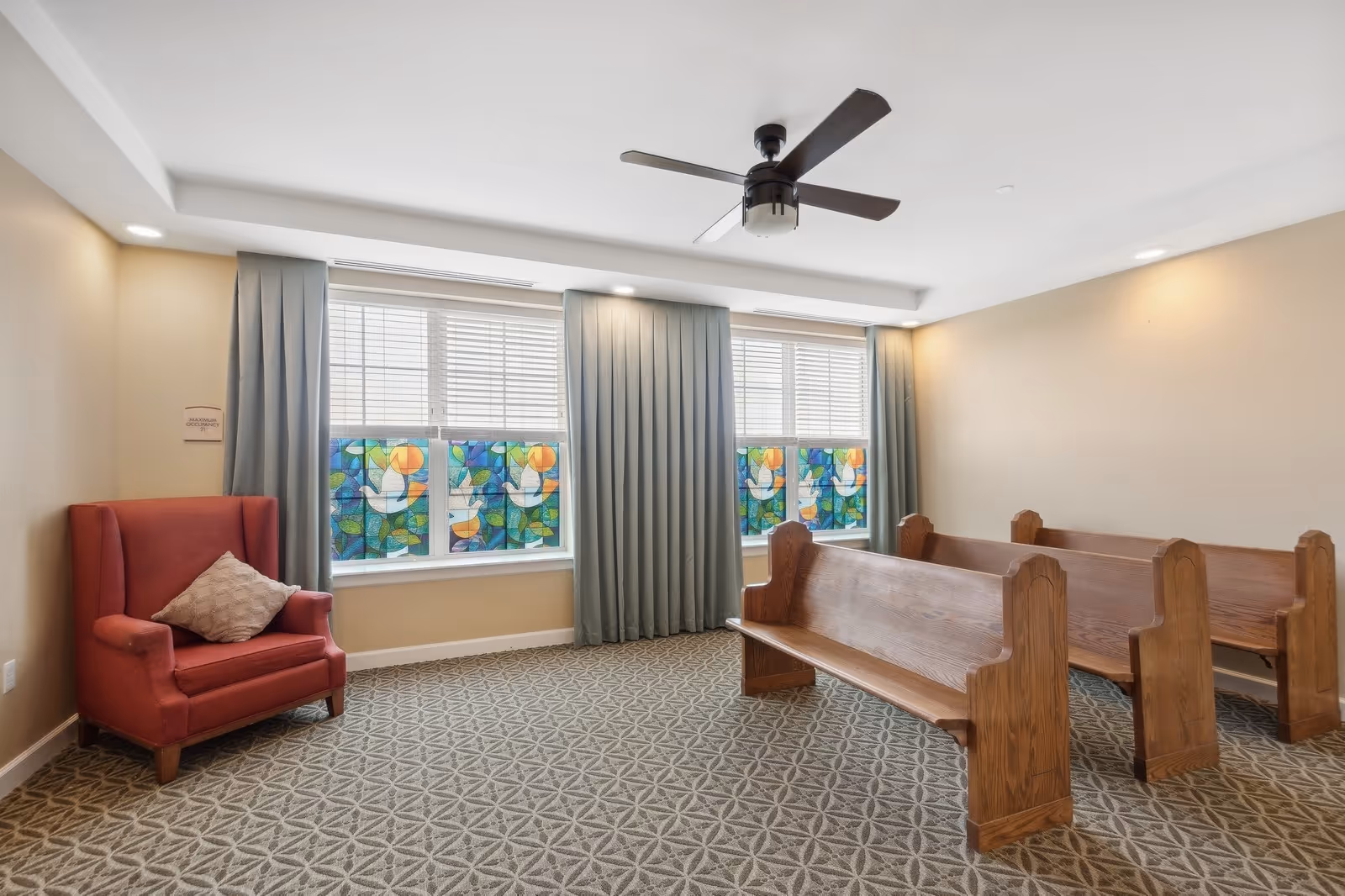 A quiet room with wooden pew-style benches arranged in rows facing a wall with large windows covered by light blue curtains and stained glass panels depicting white doves and colorful abstract shapes. There is a red upholstered armchair with a beige pillow in the corner, beige walls, patterned carpet, and a ceiling fan with dark blades.