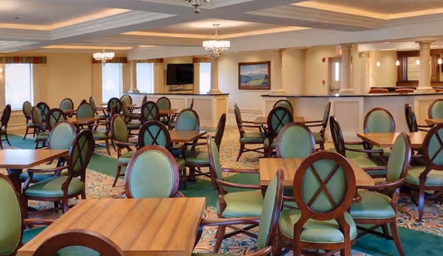 Large communal dining room with wooden tables and green upholstered chairs, chandeliers, and a TV on the far wall.
