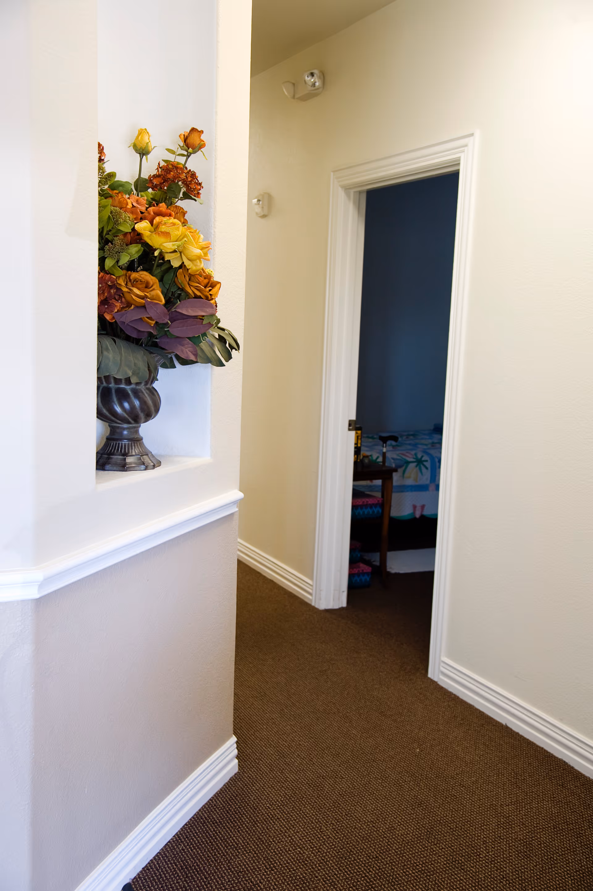 A hallway with beige walls and brown carpet leading to a bedroom. On the left side of the hallway, there is a built-in wall niche holding a decorative vase filled with orange, yellow, and purple flowers. The bedroom door is open, showing part of a bed with a colorful bedspread.