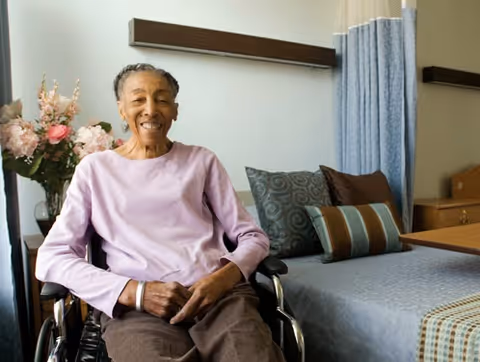 An elderly woman sitting in a wheelchair smiling in a room with a bed, pillows, and a vase of flowers on a table in the background.
