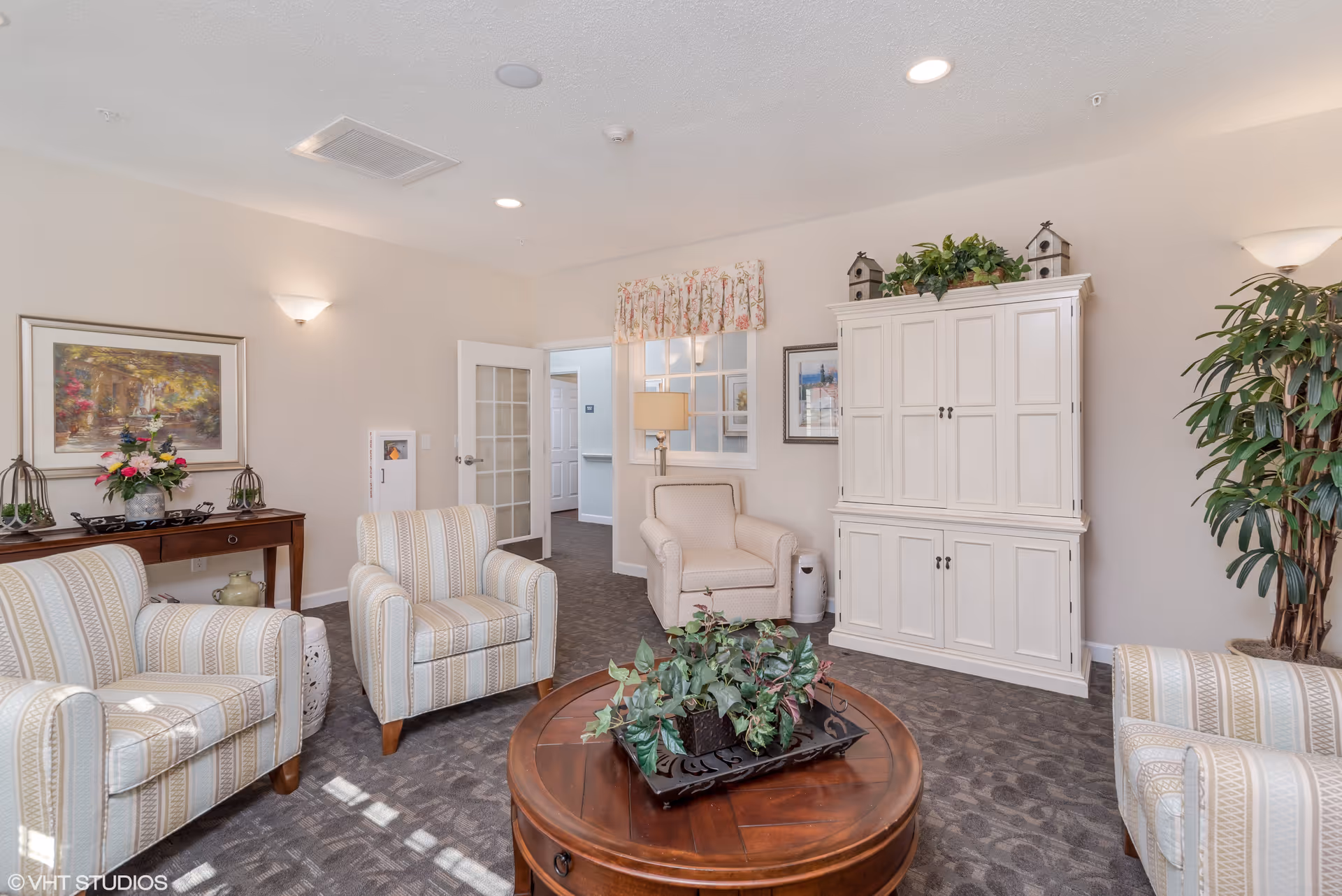 A cozy senior living common area with four striped armchairs arranged around a round wooden coffee table with a plant centerpiece. The room features a white cabinet with decorative items on top, a side table with flowers, framed artwork on the walls, a large potted plant, and soft lighting from wall sconces and a table lamp.