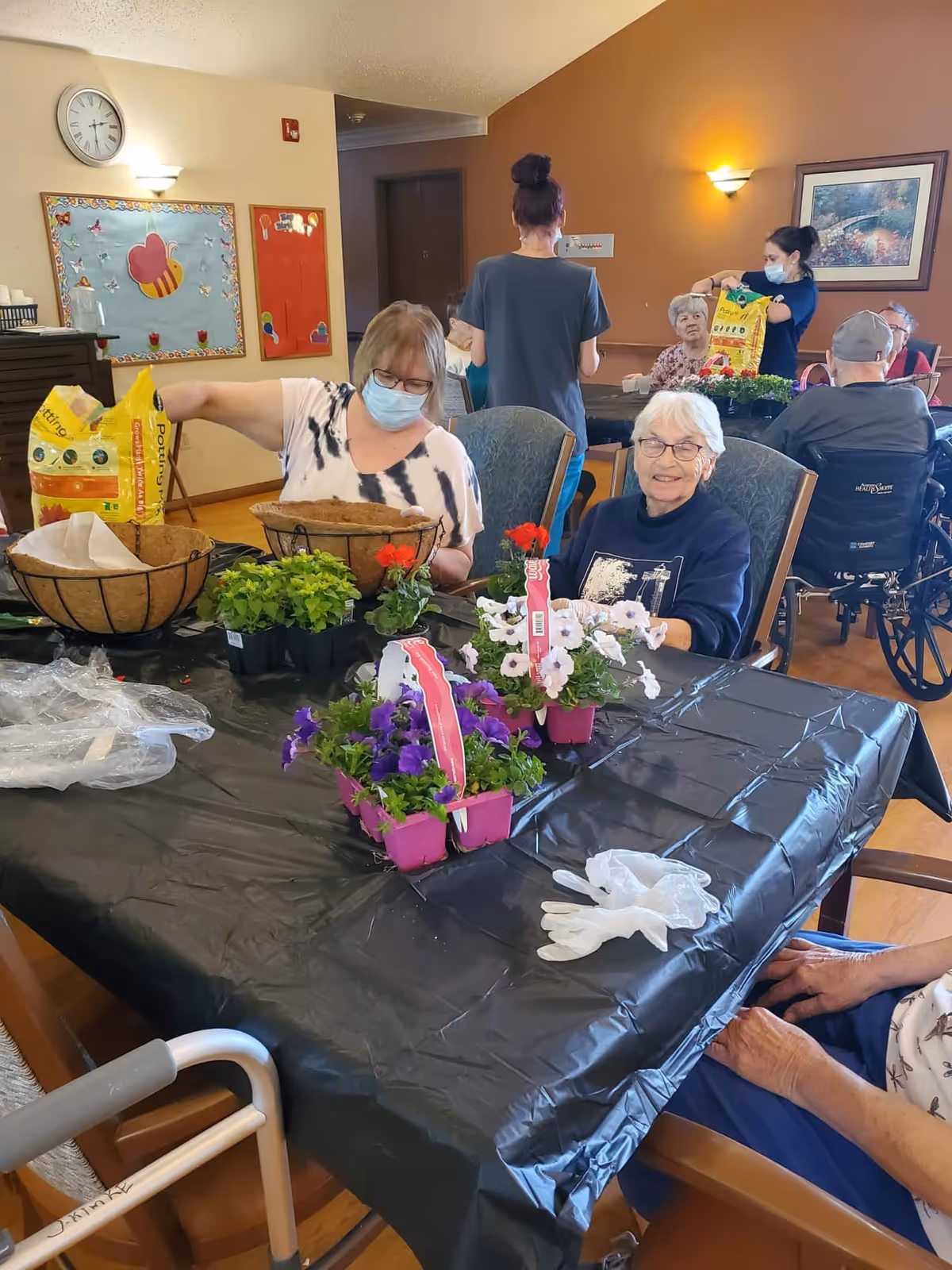 A group of elderly individuals and caregivers in a senior living facility are seated around a table covered with a black plastic tablecloth. They are engaged in a gardening activity with potted flowers and planting supplies. Some people are wearing face masks. The room has warm-colored walls, a clock, and bulletin boards in the background.