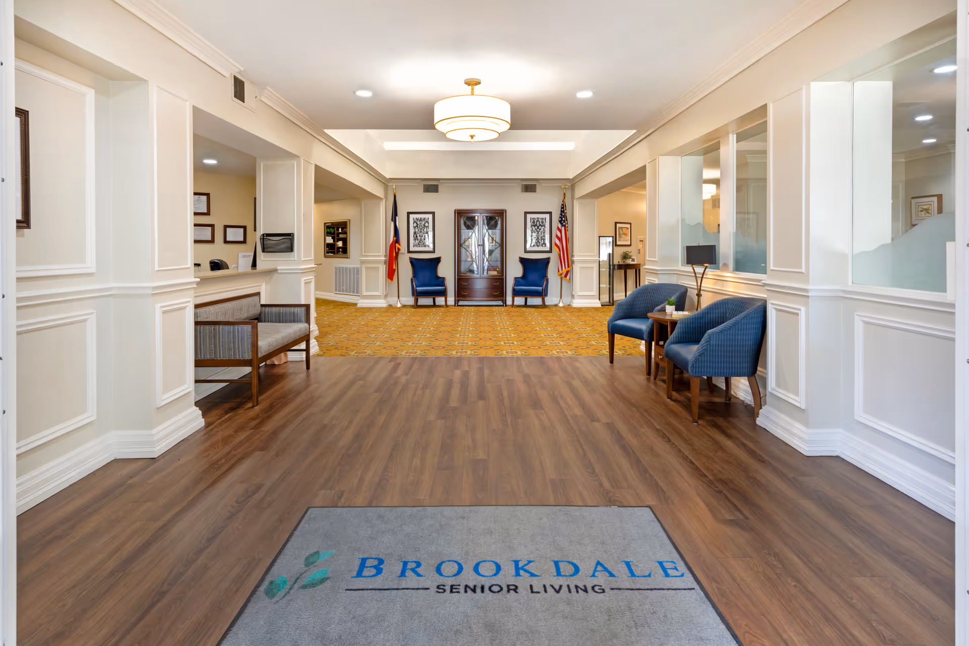 Entrance lobby of Brookdale Eden Estates senior living facility featuring a wood floor with a Brookdale Senior Living branded mat, beige walls with white trim, blue upholstered chairs, a wooden bench, a central light fixture, and flags displayed near a wooden cabinet at the far end.