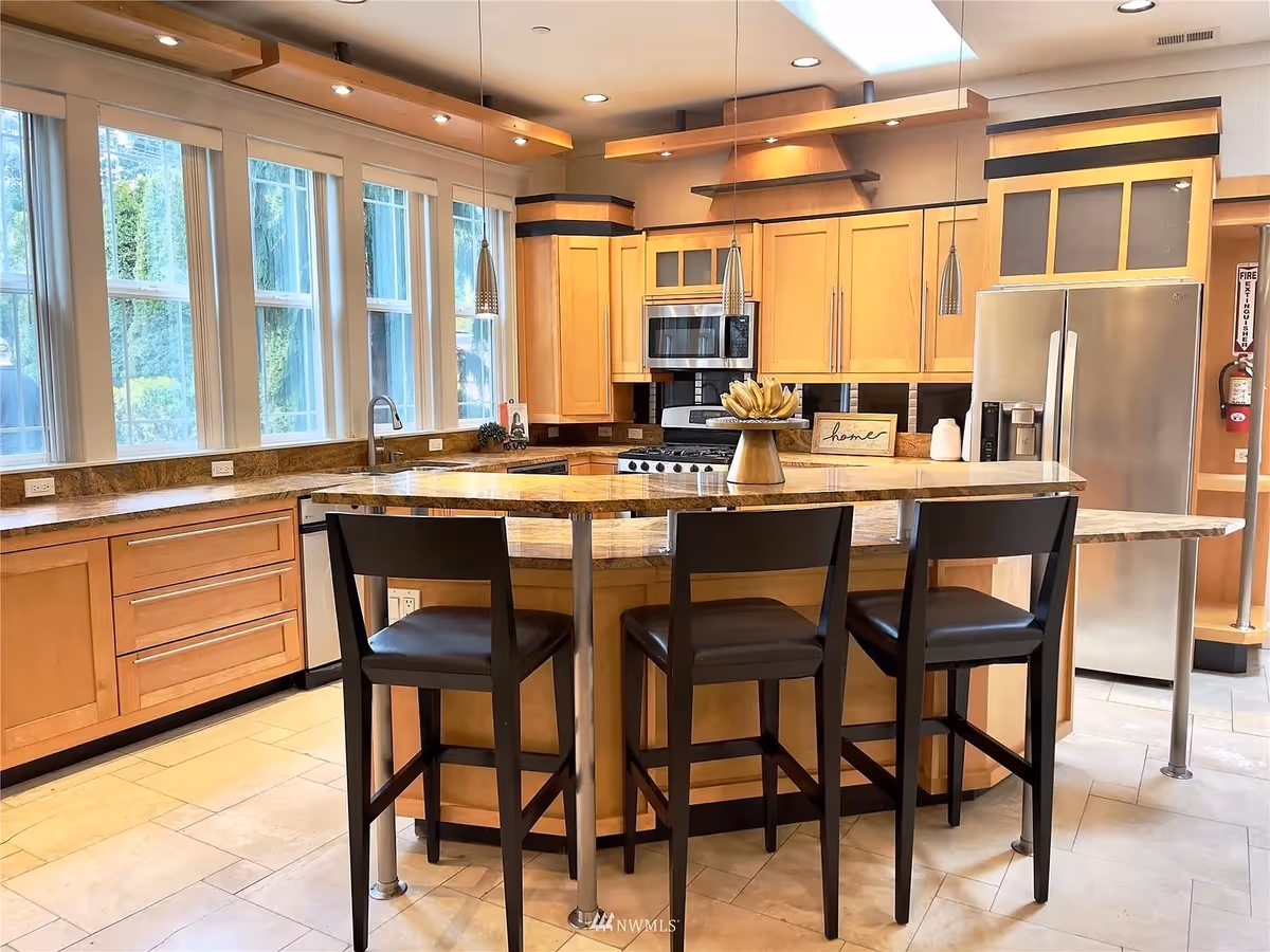 A modern kitchen with light wood cabinetry, a large island with a granite countertop, and three black bar stools. The kitchen features stainless steel appliances including a refrigerator, microwave, and stove. Large windows let in natural light, and pendant lights hang above the island. A small decorative sign that says 'home' is placed on the countertop.