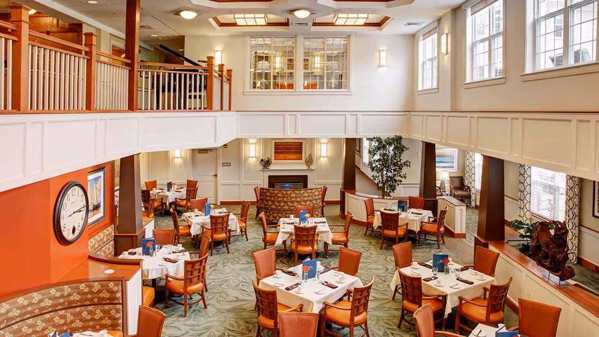 A spacious dining room with multiple tables covered in white tablecloths, each set with plates, napkins, and utensils. The room features wooden chairs with orange cushions, a large clock on an orange accent wall, a fireplace, large windows allowing natural light, and a second-floor balcony overlooking the area.
