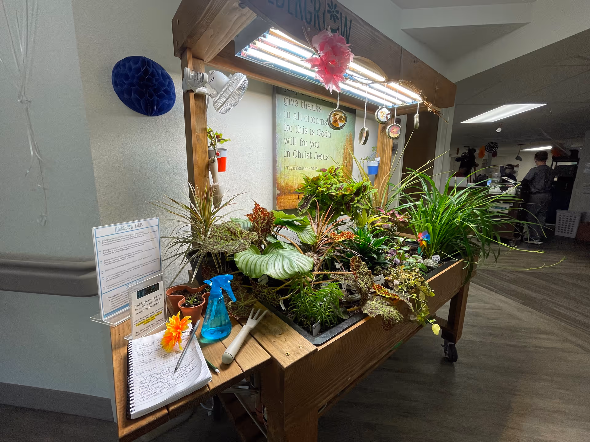 Indoor gardening station with various potted plants under grow lights on a wooden table. A blue spray bottle, small gardening tools, and a notebook with handwritten notes are on the table. A sign with instructions and a framed inspirational quote are visible on the wall behind the plants. The setting appears to be inside a senior living facility hallway with exercise equipment and people in the background.