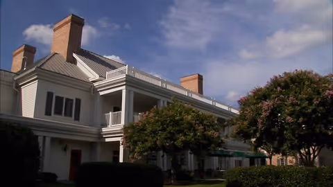Two-story retirement building with white siding, balconies, and trimmed trees under blue sky