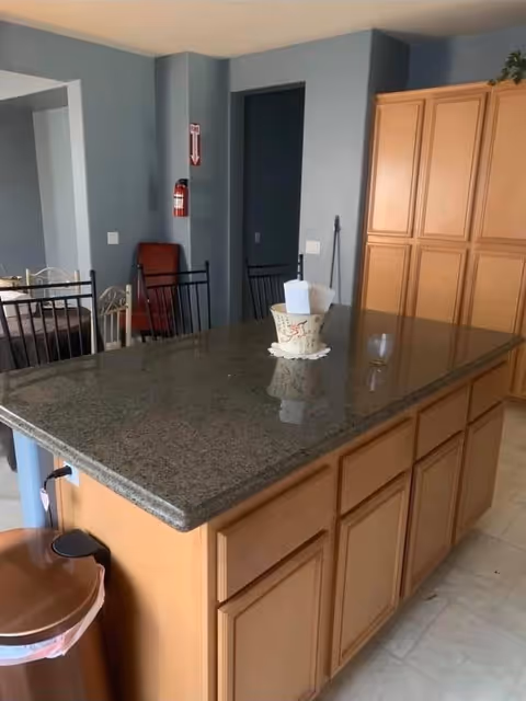 Interior view of a kitchen area featuring a large granite countertop island with wooden cabinets underneath. There are several black metal chairs around the island, a trash can with a brown lid on the side, and a decorative container with napkins on the countertop. In the background, there are more wooden cabinets, a fire extinguisher mounted on a blue-gray wall, and a doorway leading to another room.