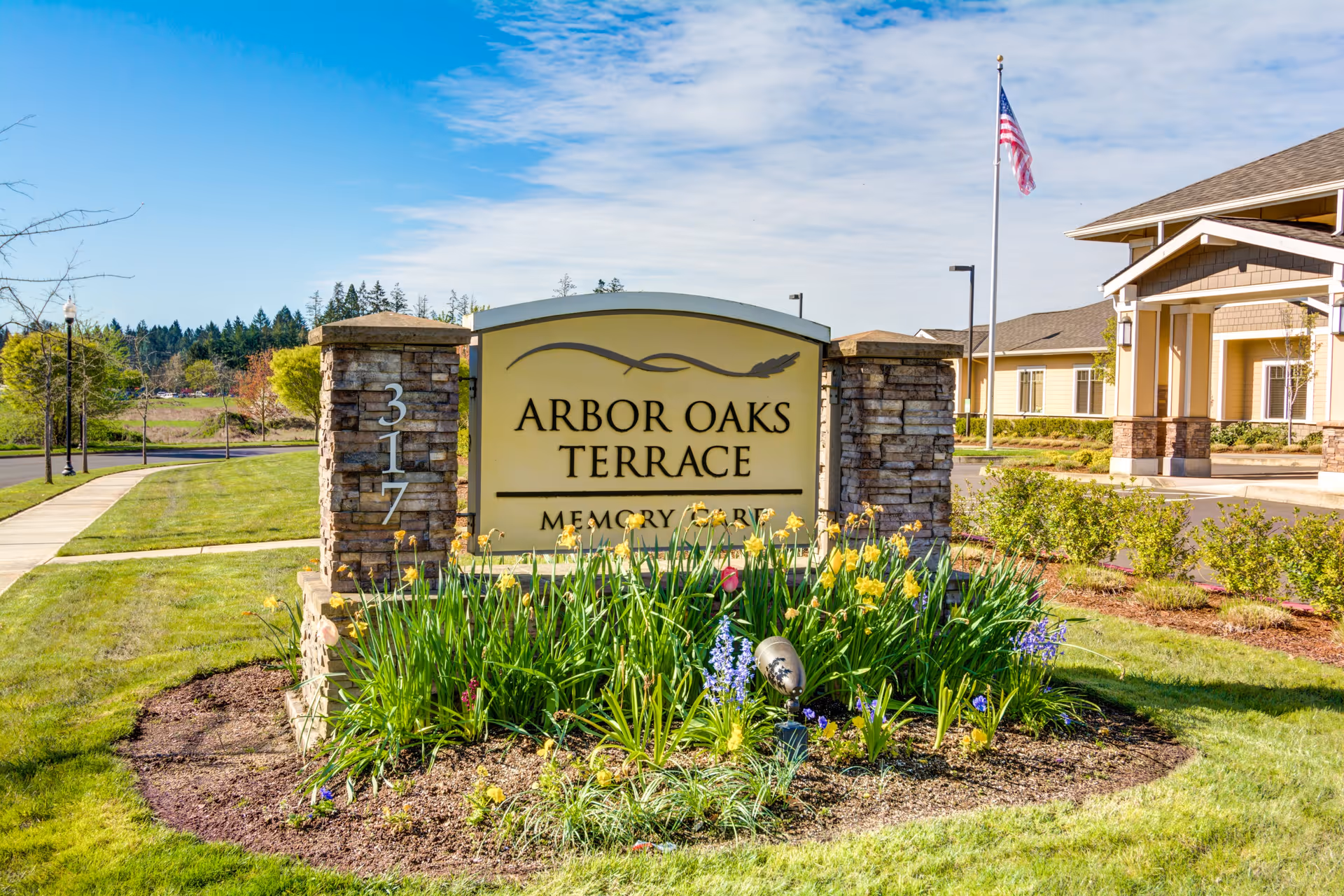 Outdoor view of the entrance sign for Arbor Oaks Terrace Memory Care Residence, surrounded by a flower bed with yellow and purple flowers, green grass, and a sidewalk leading to the building. The building and an American flag on a flagpole are visible in the background under a partly cloudy blue sky.