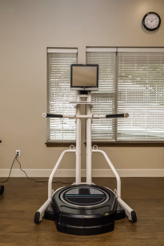 A piece of exercise equipment with a monitor screen in front of two windows with blinds. The equipment is positioned on a wooden floor against a beige wall with a clock showing the time as 12:15.