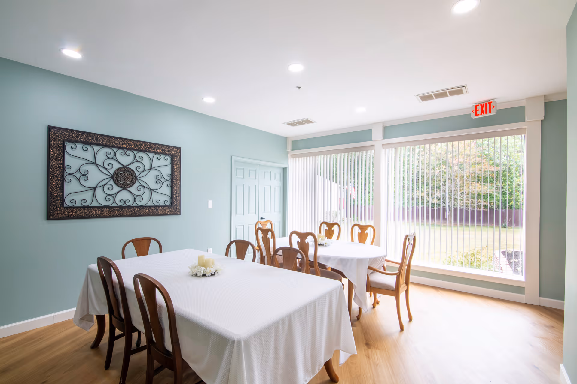 A bright dining room with two tables covered in white tablecloths, surrounded by wooden chairs. The room has light green walls, a large decorative wrought iron wall art, and a large window with vertical blinds letting in natural light. The floor is wooden, and there is an exit sign above the window.