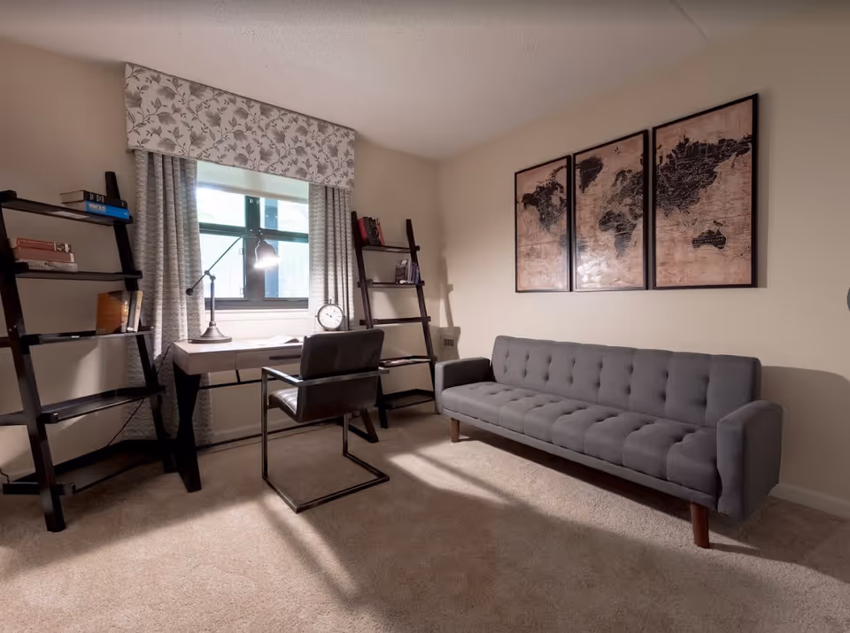 A cozy room with a gray tufted sofa against a beige wall, three framed world map prints above it, a desk with a black chair in front of a window with patterned curtains, and two dark wooden ladder-style bookshelves on either side of the desk holding books and decorative items.