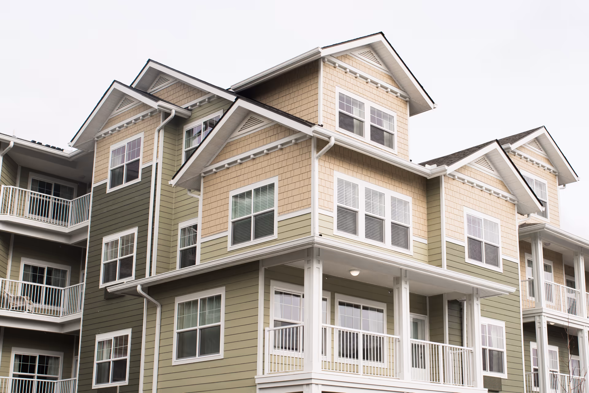 Exterior view of a multi-story residential building with beige and green siding, multiple windows, and balconies with white railings under an overcast sky.