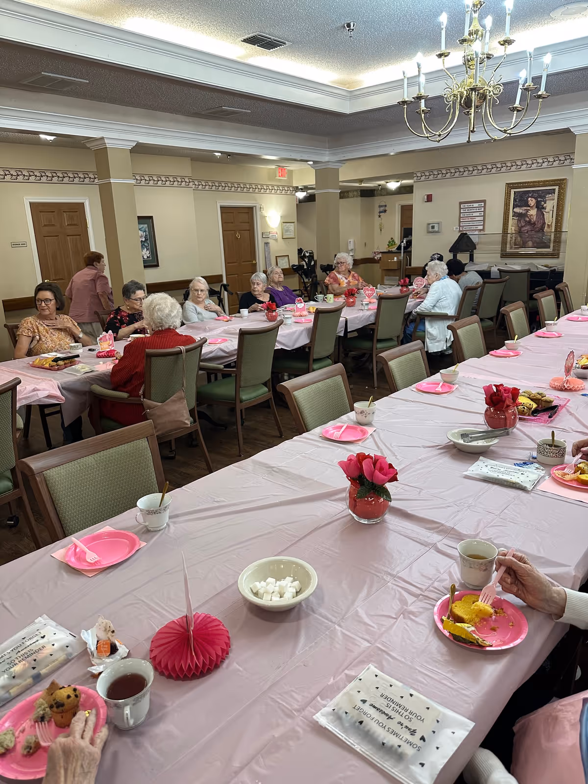 A group of elderly women seated around a long table covered with a pink tablecloth in a dining room. The table is set with pink plates, cups, and small decorations including red flowers. The room has beige walls, wooden doors, and framed artwork. Some women are eating while others are engaged in conversation.