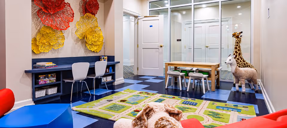 A colorful children's playroom with a patterned play mat on the floor, a small wooden table with four chairs, two large stuffed animals (a giraffe and a llama), a blue desk with two white chairs, and large decorative wall art in red and yellow resembling flowers.