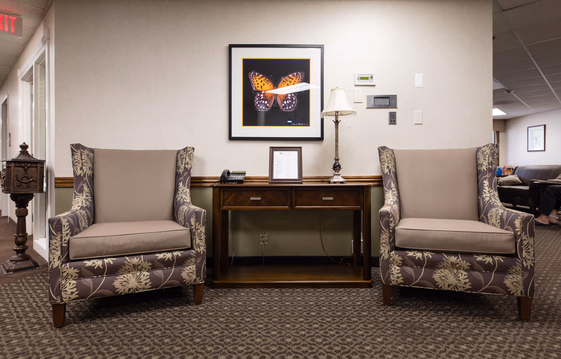 Two patterned armchairs flank a wooden console table with a lamp and framed butterfly artwork in a facility seating area.