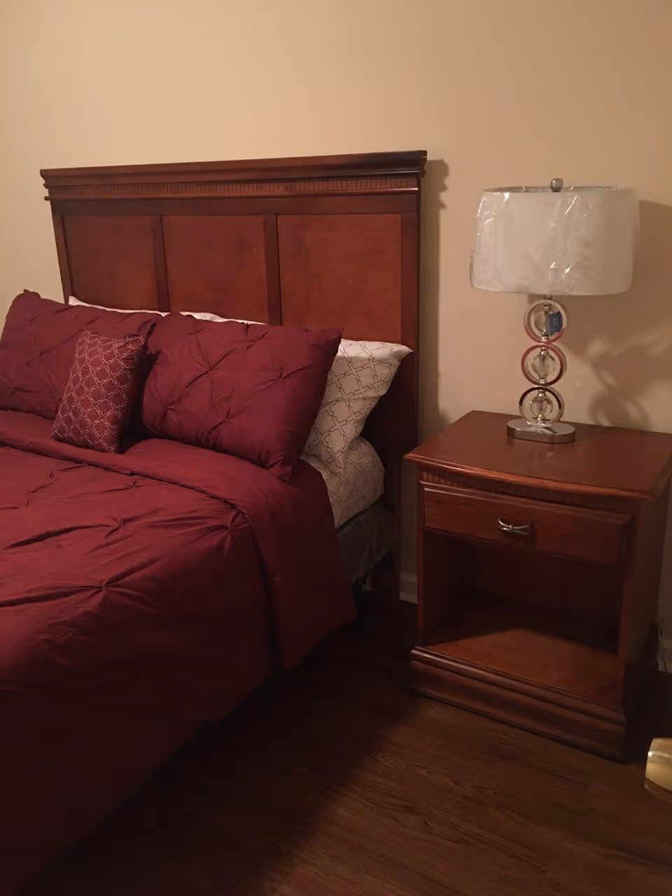 A bedroom with a wooden headboard bed dressed in burgundy bedding and pillows, next to a wooden nightstand with a modern lamp on top. The room has beige walls and wooden flooring.