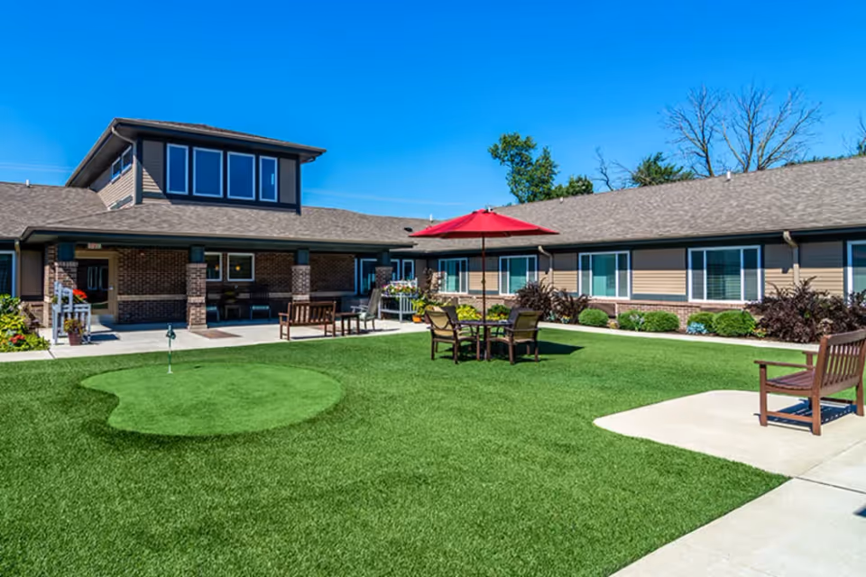 Outdoor courtyard area at Porter Place featuring a putting green, patio table with chairs and a red umbrella, benches, and a building with multiple windows under a clear blue sky.