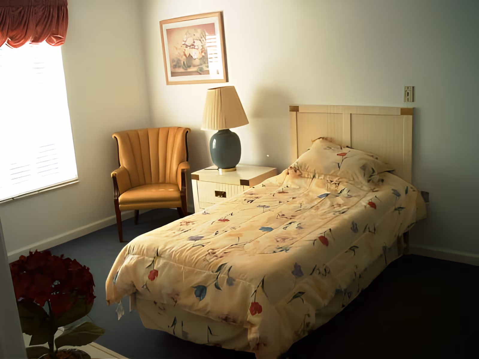 A bedroom with a single bed covered in a floral patterned comforter, a beige armchair, a nightstand with a lamp, a framed picture on the wall, and a window with a red valance curtain.