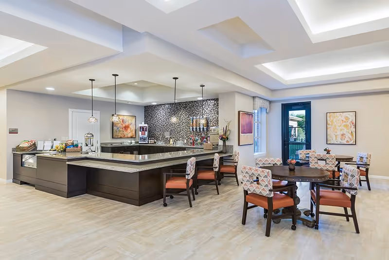 A bright and modern dining area with a large U-shaped counter featuring a granite countertop and several pendant lights hanging above. The room has multiple round wooden tables surrounded by cushioned chairs with patterned upholstery and orange seats. The walls are decorated with framed artwork, and there is a door leading outside with a window beside it. The floor is light-colored wood, and the ceiling has recessed lighting and a coffered design.