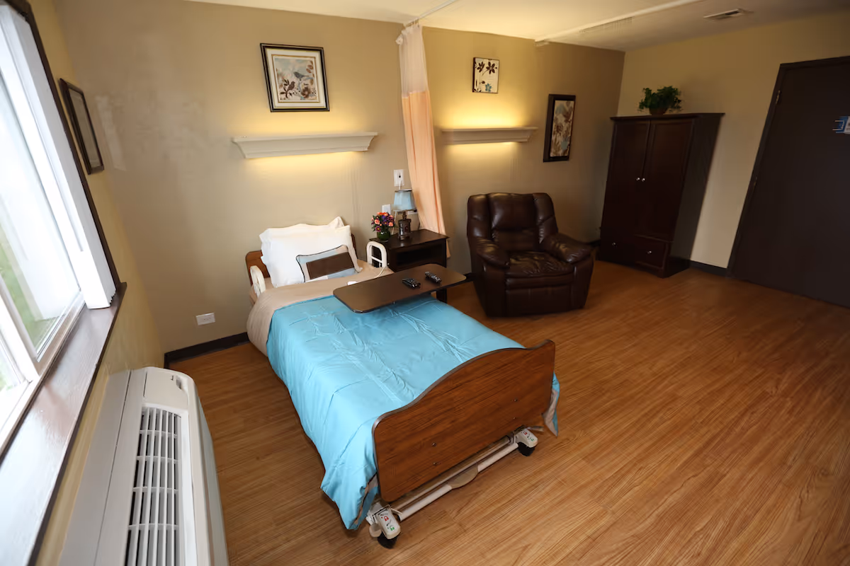A senior care patient room with a single bed covered in a blue blanket, a leather recliner, bedside table and wooden cabinet.