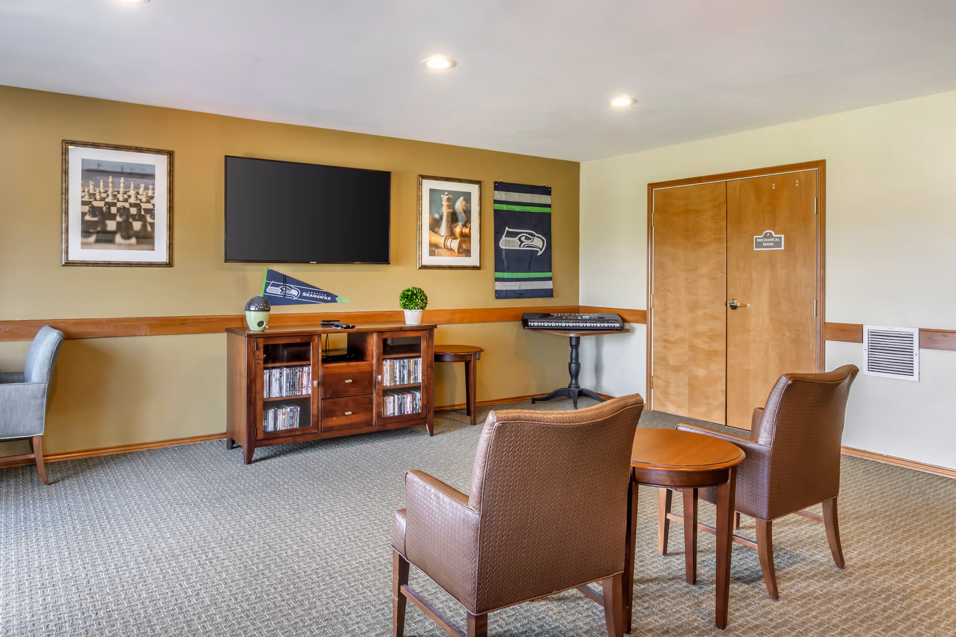 A furnished common room with two armchairs and a small round table facing a wall-mounted TV and wooden media console.