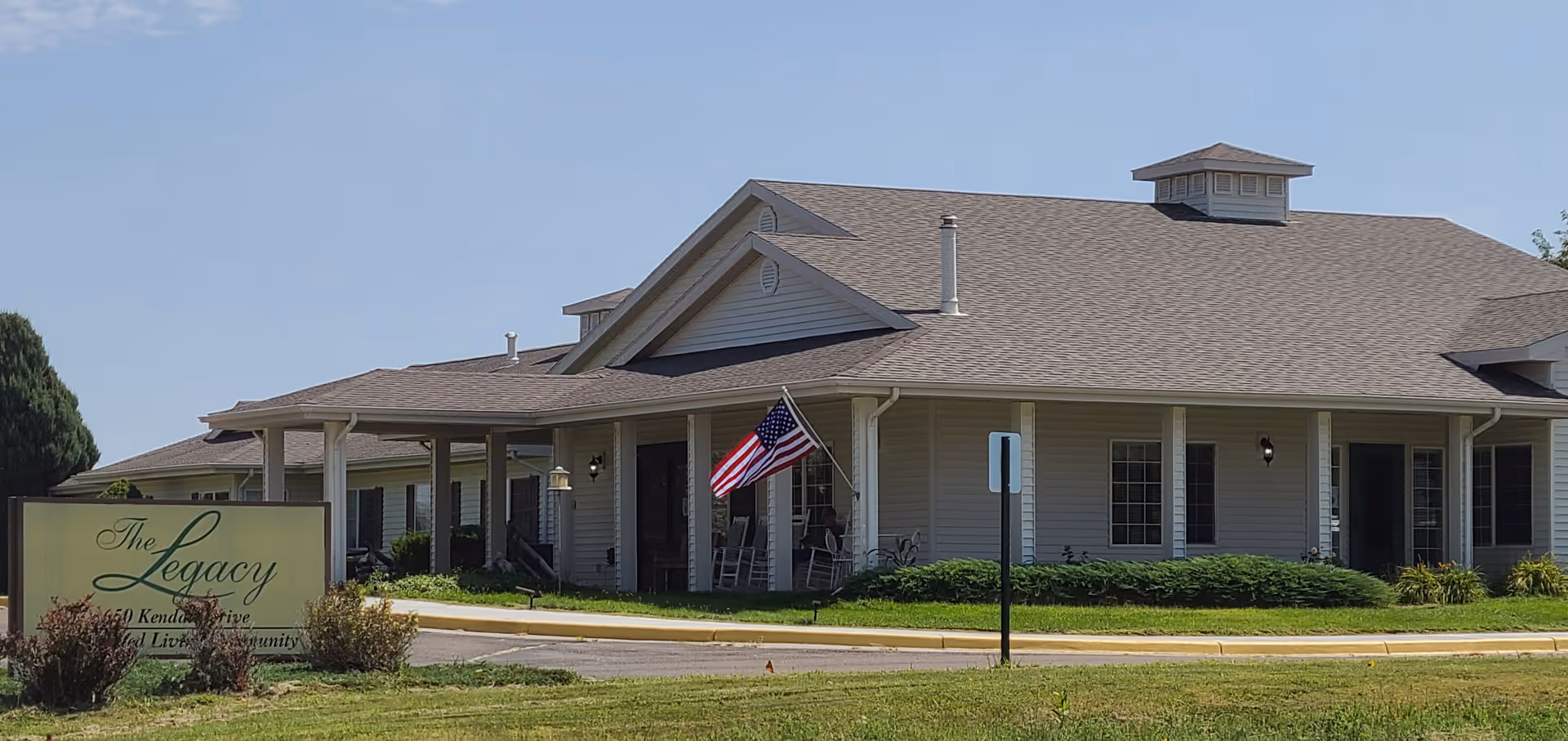 Single-story senior living building with a covered front porch, an American flag, and a sign reading 'The Legacy' in front on a sunny day.
