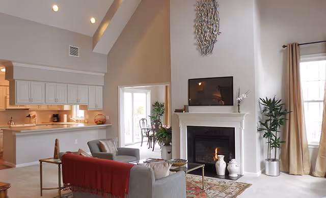 A bright and spacious living room with high vaulted ceilings, a fireplace with a mounted TV above it, a gray sofa with a red throw blanket, a glass coffee table, and large windows with beige curtains. The room opens into a kitchen area with white cabinets and a breakfast bar, and a dining area with a table and chairs is visible through a doorway.
