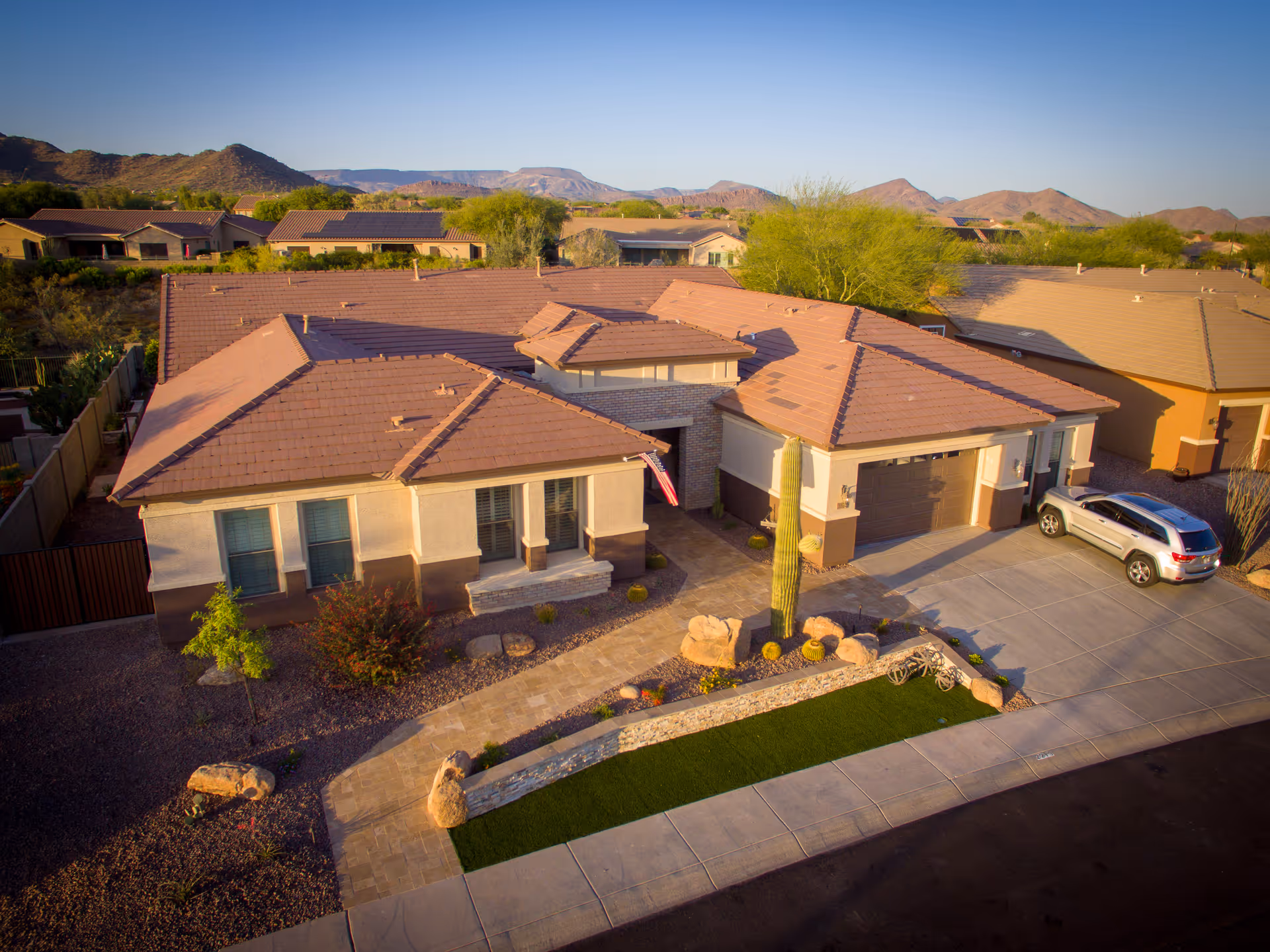 Aerial view of a single-story residential building with a tiled roof, a driveway with a parked silver SUV, desert landscaping including a tall cactus and rocks, and surrounding houses with mountains in the background under a clear sky.
