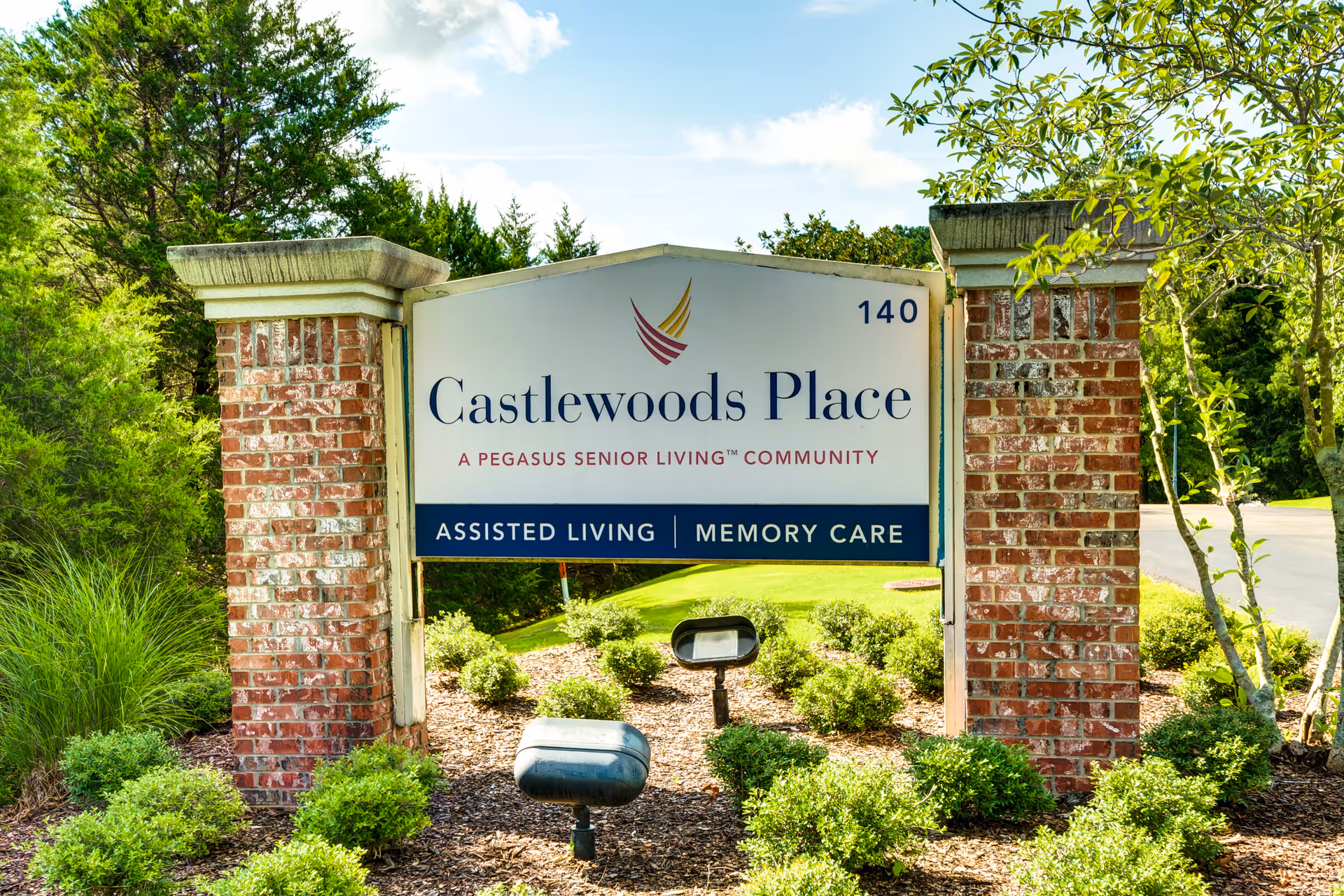 Outdoor sign for Castlewoods Place, a Pegasus Senior Living community offering assisted living and memory care, mounted between two brick pillars with greenery and trees in the background.