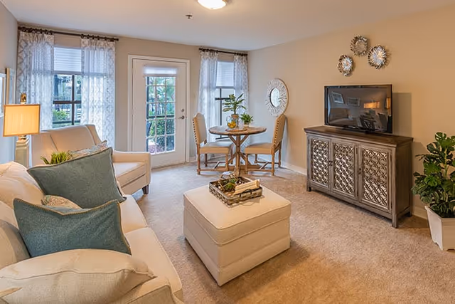 A cozy living room with beige carpet and walls, featuring a cream-colored sofa with blue and beige pillows, a matching armchair, and a square ottoman in the center. A small round wooden table with two chairs is placed near a glass door and windows with sheer curtains. A flat-screen TV sits on a decorative wooden cabinet against the wall, accompanied by wall decor and a potted plant.
