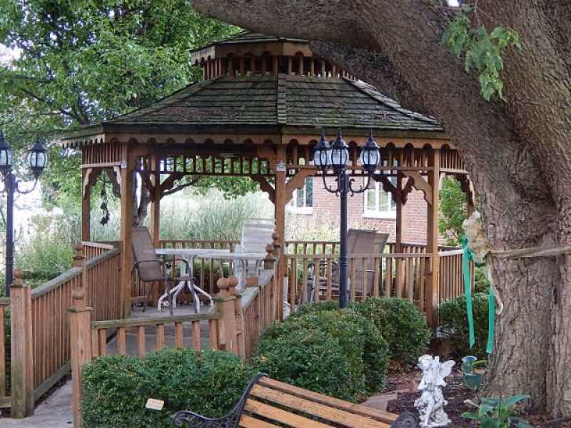 A wooden gazebo with a shingled roof surrounded by green bushes and trees. Inside the gazebo are several chairs and a table. A black vintage-style street lamp with three lanterns stands near the gazebo. In the foreground, there is a wooden bench and a small angel statue near the base of a large tree.