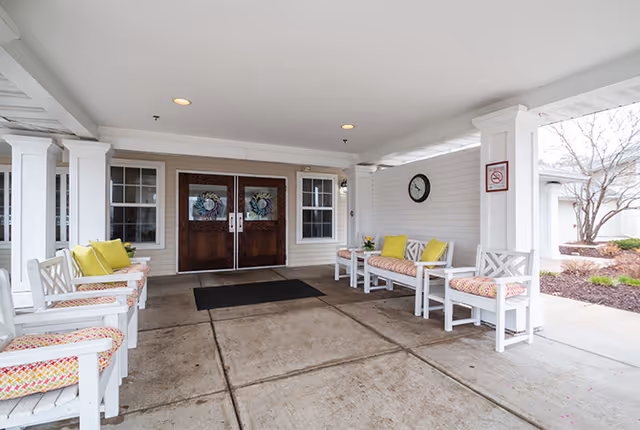 Covered entrance area of a senior living facility with white wooden chairs and benches featuring colorful cushions and pillows. Double wooden doors with decorative wreaths are centered at the back, flanked by windows. A clock and a no smoking sign are mounted on the white wall to the right. The floor is concrete and the area is well-lit with ceiling lights.