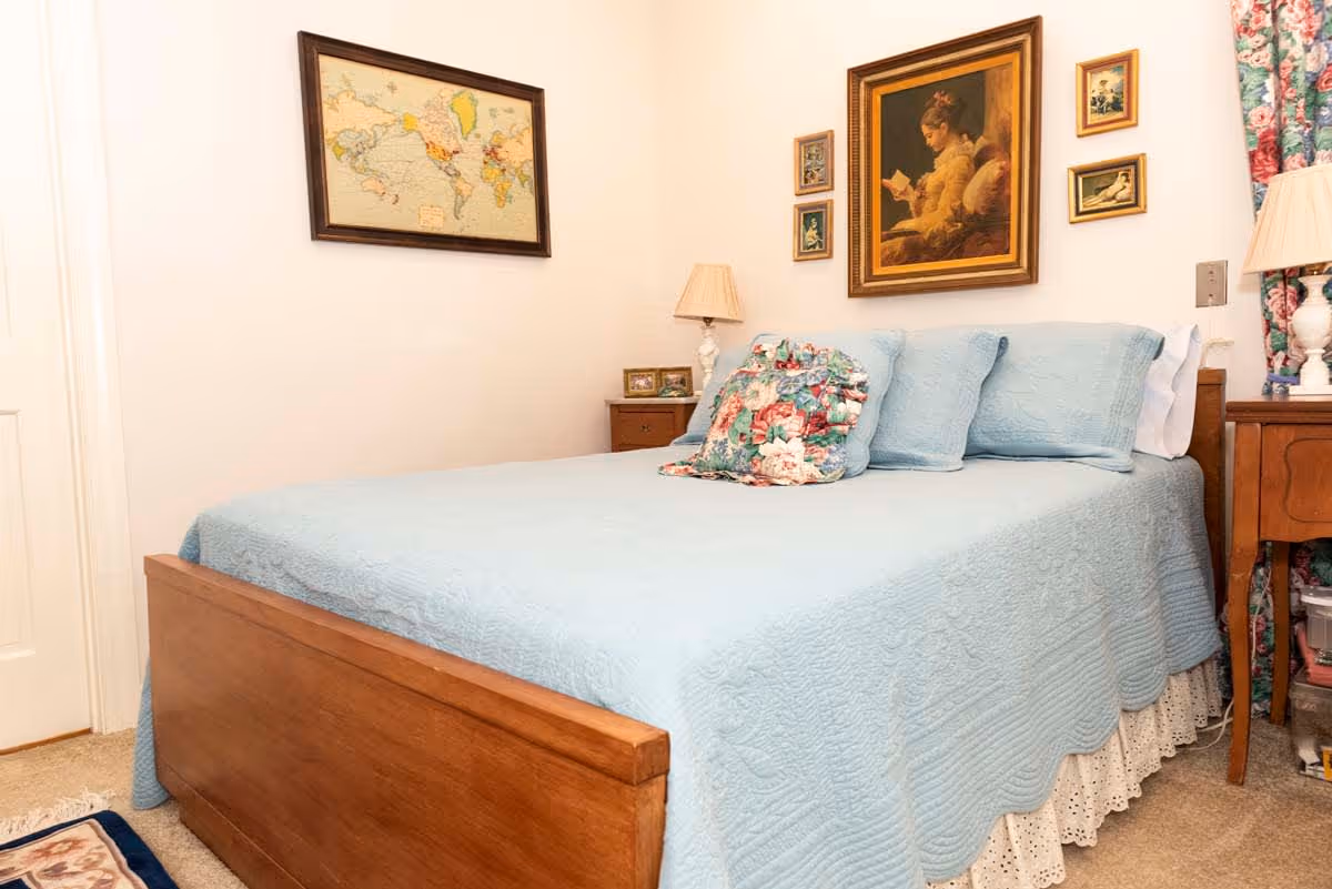 A cozy bedroom with a wooden bed frame and a light blue quilted bedspread. The bed has matching blue pillows and a floral decorative pillow. On the wall above the bed are several framed pictures, including a large painting of a girl reading and a world map. There are two wooden nightstands on either side of the bed, each with a lamp. Floral curtains are partially visible on the right side of the image.